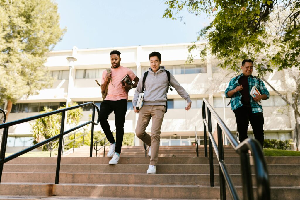 pexels-photo-7683750-7683750 Three college students walk down stairs on campus, carrying books and backpacks, on a sunny day.